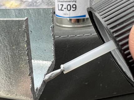 En person påfører omhyggeligt en væske fra en lille flaske på en metaloverflade, hvilket demonstrerer præcisionen ved at bruge "Zinkreparation med Tyk Belægning til Galvaniserede Overflader – 100 ml Sølvlys." I baggrunden foreslår en dåse mærket "ReiColor LZ-09" brugen af avancerede restaureringsmaterialer. De udendørs omgivelser tilbyder et ideelt miljø til denne omhyggelige opgave.