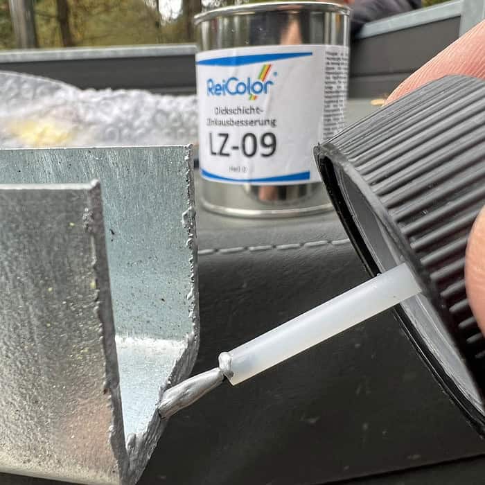 En person påfører omhyggeligt en væske fra en lille flaske på en metaloverflade, hvilket demonstrerer præcisionen ved at bruge "Zinkreparation med Tyk Belægning til Galvaniserede Overflader – 100 ml Sølvlys." I baggrunden foreslår en dåse mærket "ReiColor LZ-09" brugen af avancerede restaureringsmaterialer. De udendørs omgivelser tilbyder et ideelt miljø til denne omhyggelige opgave.