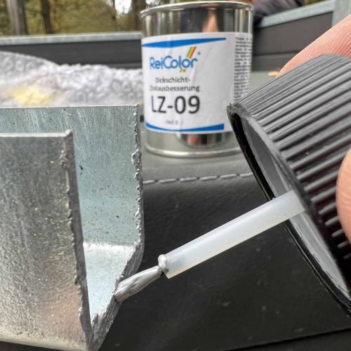 En person påfører omhyggeligt en væske fra en lille flaske på en metaloverflade, hvilket demonstrerer præcisionen ved at bruge "Zinkreparation med Tyk Belægning til Galvaniserede Overflader – 100 ml Sølvlys." I baggrunden foreslår en dåse mærket "ReiColor LZ-09" brugen af avancerede restaureringsmaterialer. De udendørs omgivelser tilbyder et ideelt miljø til denne omhyggelige opgave.