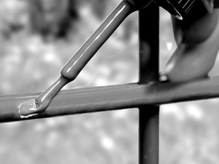 En hånd, der holder en pensel, påfører et nyt lag maling på et metalhegn ved hjælp af Reparationskit til Metal – Sort RAL 9005 (20 ml Penselflaske). Penslen er vist tæt på, med Sort RAL 9005 maling, der synligt dækker hegnsstængerne. Baggrunden er sløret og fokuserer opmærksomheden på maleraktiviteten. Billedet er i sort/hvid.