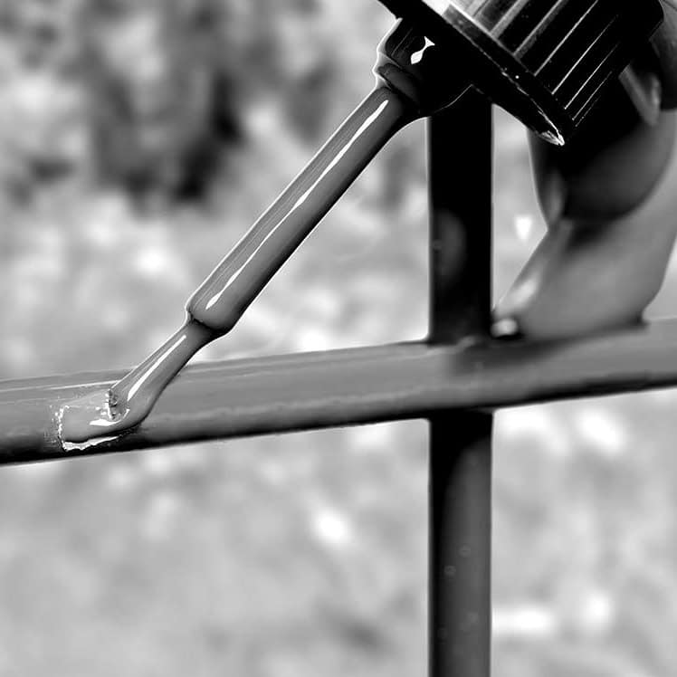 En hånd, der holder en pensel, påfører et nyt lag maling på et metalhegn ved hjælp af Reparationskit til Metal – Sort RAL 9005 (20 ml Penselflaske). Penslen er vist tæt på, med Sort RAL 9005 maling, der synligt dækker hegnsstængerne. Baggrunden er sløret og fokuserer opmærksomheden på maleraktiviteten. Billedet er i sort/hvid.