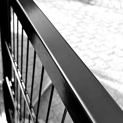 Sort/hvid billede af et rækværk. Fokus er på rækværkets vandrette og lodrette bjælker med en sløret, brostenslignende overflade i baggrunden. Billedet fremhæver den glatte, reflekterende overflade af metallet.