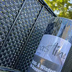 BASTLINE Afskærmning rulle 26m Polyrattan til Panelhegn er udstillet mod et hegn i fletstil, med dets ternede mønster, der forstærker baggrundens tekstur. Sollyset filtrerer blødt gennem det nærliggende grønne område og skaber en lys og rolig atmosfære, der yderligere fremhæves af polyrattan-designet.