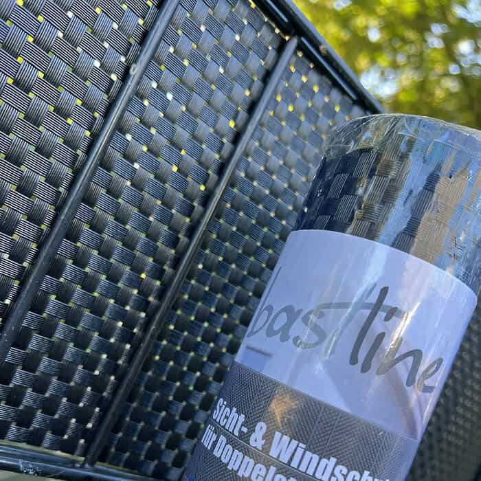 BASTLINE Afskærmning rulle 26m Polyrattan til Panelhegn er udstillet mod et hegn i fletstil, med dets ternede mønster, der forstærker baggrundens tekstur. Sollyset filtrerer blødt gennem det nærliggende grønne område og skaber en lys og rolig atmosfære, der yderligere fremhæves af polyrattan-designet.