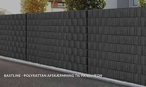 Et sort polyrattan hegn langs en forstadsgade, hvilket giver privatliv. En slank stribe græs løber langs barrieren, med træer synlige i baggrunden. Tekst på dansk lyder: "BASTLINE Afskærmning rulle 26m Polyrattan til Panelhegn.