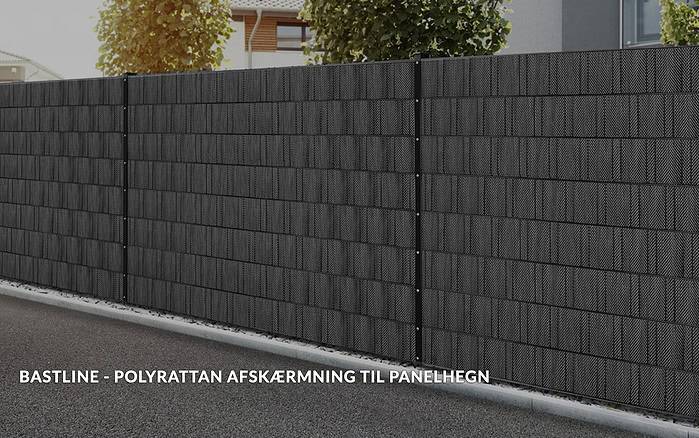 Et sort polyrattan hegn langs en forstadsgade, hvilket giver privatliv. En slank stribe græs løber langs barrieren, med træer synlige i baggrunden. Tekst på dansk lyder: "BASTLINE Afskærmning rulle 26m Polyrattan til Panelhegn.
