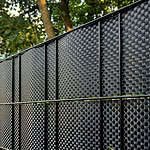 Nærbillede af en BASTLINE Afskærmning rulle 26m Polyrattan til Panelhegn i sort, monteret under et træ. Sollyset filtrerer gennem bladene og kaster plettede skygger på hegnets strukturerede overflade.