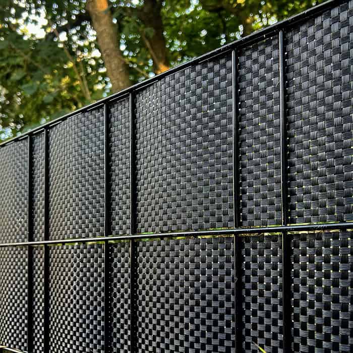 Nærbillede af en BASTLINE Afskærmning rulle 26m Polyrattan til Panelhegn i sort, monteret under et træ. Sollyset filtrerer gennem bladene og kaster plettede skygger på hegnets strukturerede overflade.