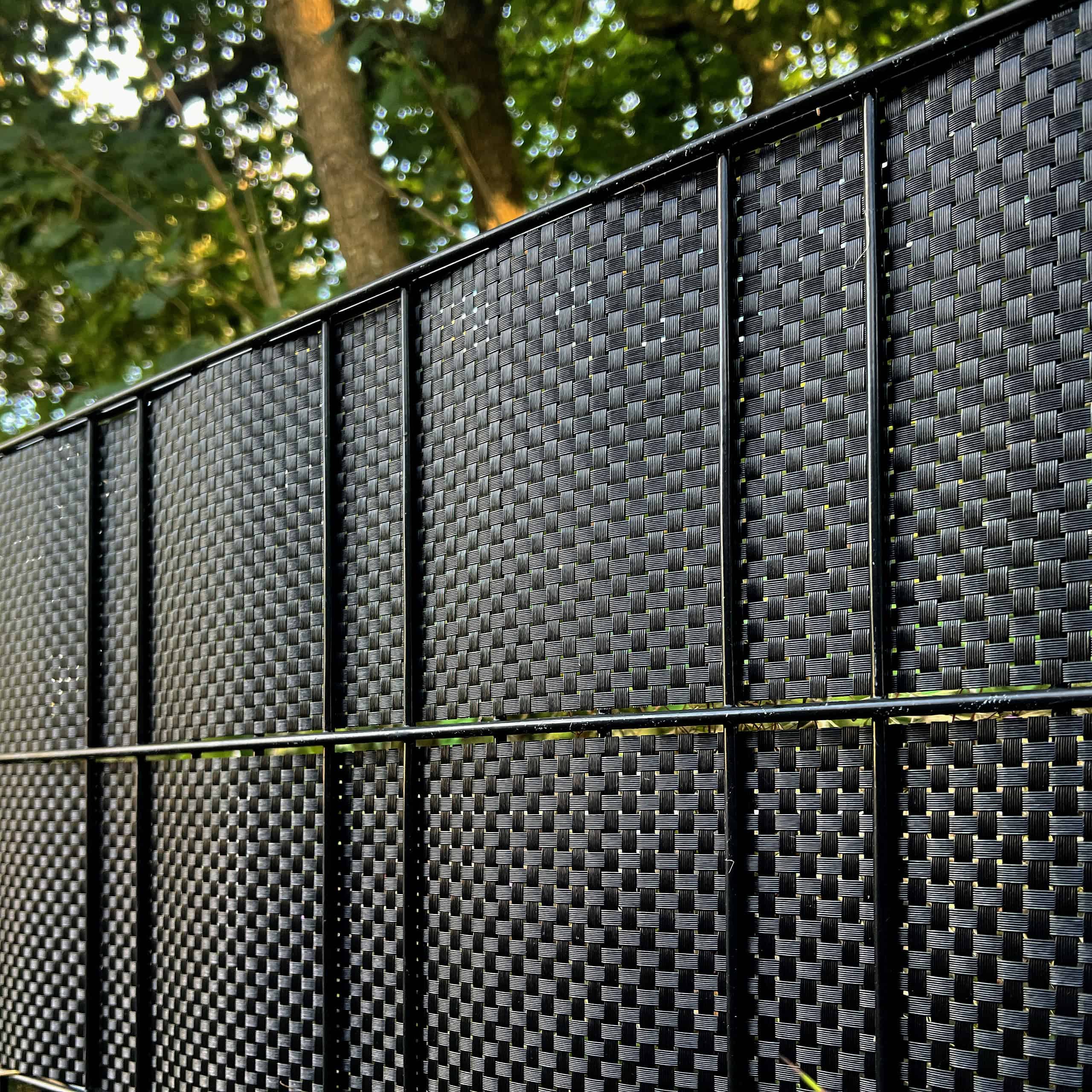 Nærbillede af en BASTLINE Afskærmning rulle 26m Polyrattan til Panelhegn i sort, monteret under et træ. Sollyset filtrerer gennem bladene og kaster plettede skygger på hegnets strukturerede overflade.
