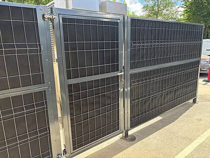Støjhegnslåge ECO 18dB – Enkeltlåge - Antracit/Varmegalvaniseret, en høj metalport med gittermønster og lås, er monteret på et matchende hegn og kaster skygger på fortovet udenfor under en solrig blå himmel med træer i baggrunden.