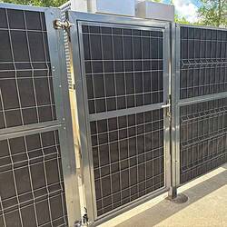 Støjhegnslåge ECO 18dB – Enkeltlåge - Antracit/Varmegalvaniseret, en høj metalport med gittermønster og lås, er monteret på et matchende hegn og kaster skygger på fortovet udenfor under en solrig blå himmel med træer i baggrunden.