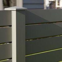 Nærbillede af VIDUAL Line – Moderne Aluminiumshegn med Spalteprofiler, der viser et gråt lamelhegn med en lodret støttestolpe. Det slanke design kaster skygger under sollys og fremhæver de vandrette linjer. Bygninger og træer er slørede i baggrunden.