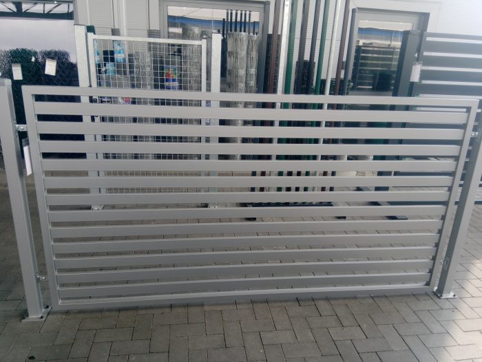 Et moderne Metal Hegn – JALOUSI hegn med vandrette lameller, pulverlakeret og vedligeholdelsesfrit, fremvises på en brolagt overflade. Bagved pryder forskellige andre hegns- og portmuligheder showroomets udstillingsområde.