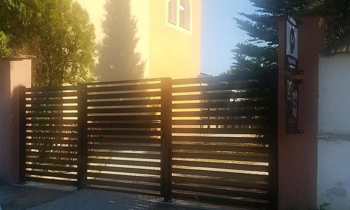 En moderne, sort Metal Hegn – JALOUSI port fremhæver den beige bygning og kaster dansende skygger i sollys. Træer og buske tilføjer et naturligt præg, mens et skilt ved siden af denne stilfulde, vedligeholdelsesfrie låge forstærker scenens charme.