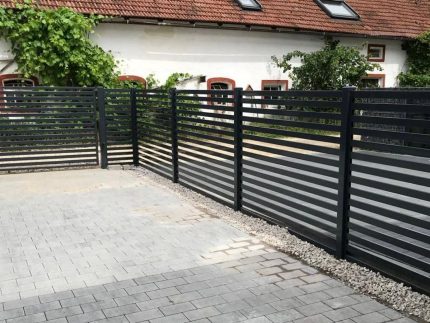 En sort Metal Hegn – JALOUSI, pulverlakeret og vedligeholdelsesfri, omslutter et asfalteret område. Grunden har grå belægningssten med en grusstrimmel langs hegnet. I baggrunden står en hvid bygning med rødindrammede vinduer og rødt tegltag.