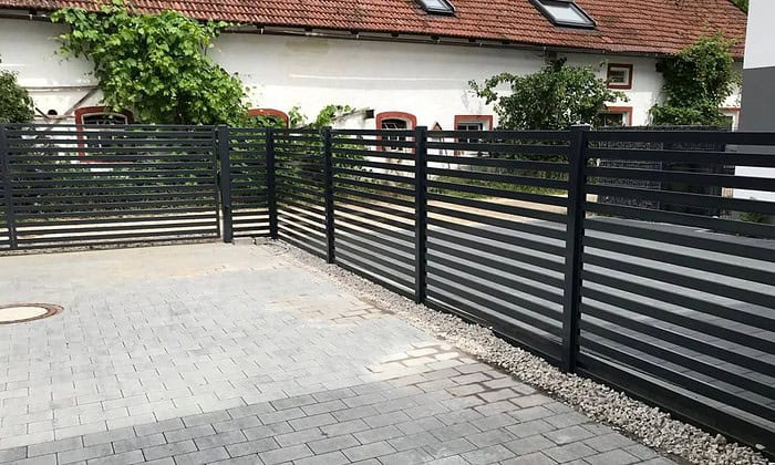 En sort Metal Hegn – JALOUSI, pulverlakeret og vedligeholdelsesfri, omslutter et asfalteret område. Grunden har grå belægningssten med en grusstrimmel langs hegnet. I baggrunden står en hvid bygning med rødindrammede vinduer og rødt tegltag.