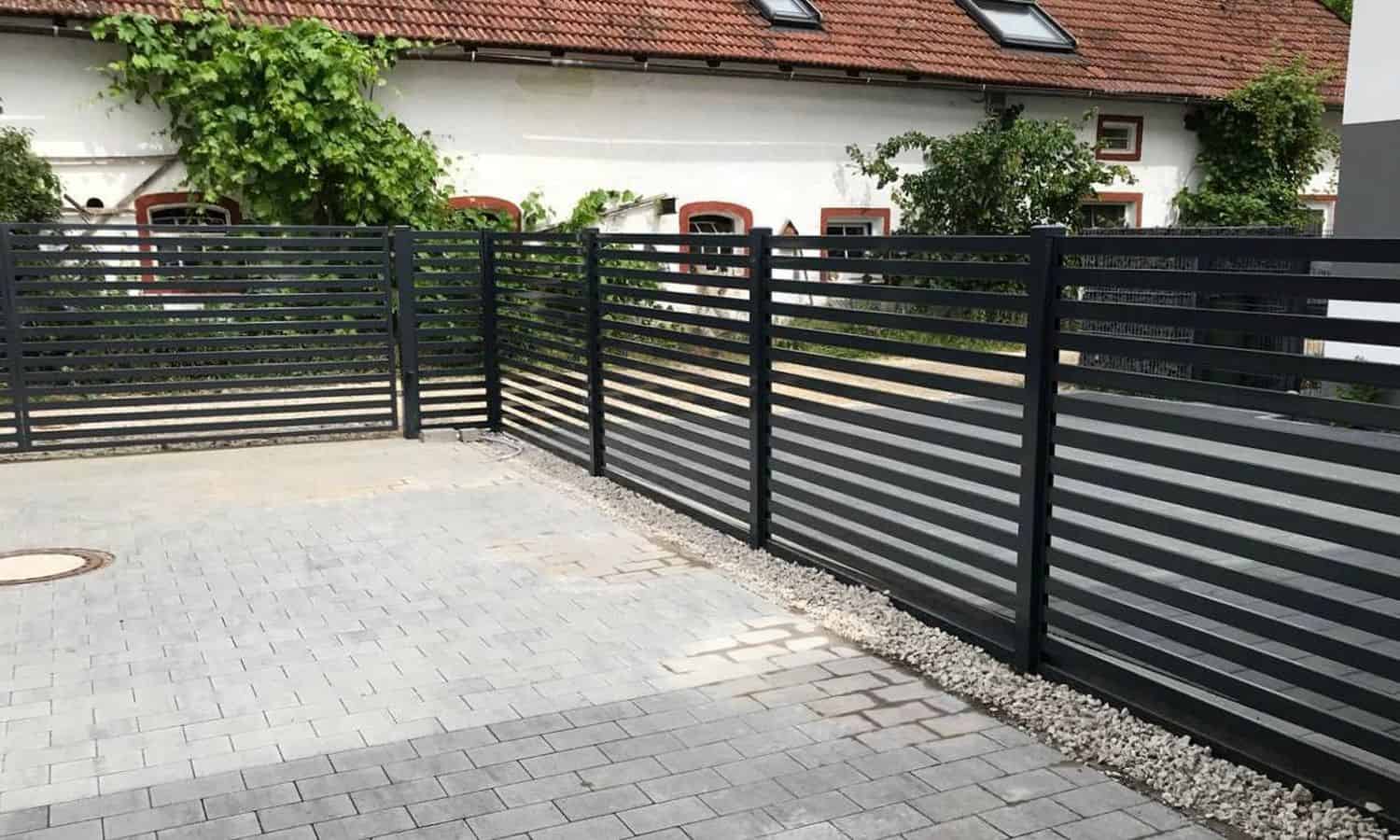 En sort Metal Hegn – JALOUSI, pulverlakeret og vedligeholdelsesfri, omslutter et asfalteret område. Grunden har grå belægningssten med en grusstrimmel langs hegnet. I baggrunden står en hvid bygning med rødindrammede vinduer og rødt tegltag.