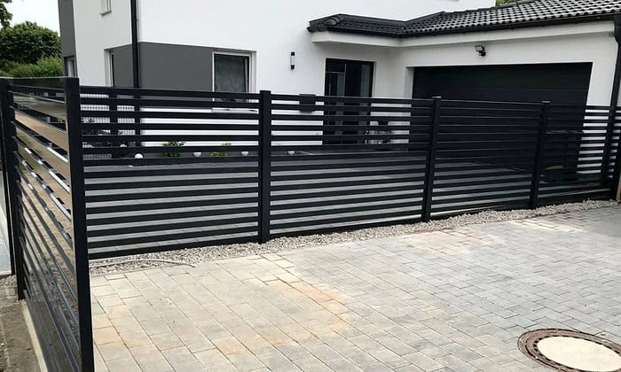 Et moderne hus med hvide vægge og sort garageport er omkranset af den slanke Metal Hegn – JALOUSI, som er pulverlakeret og vedligeholdelsesfrit. Det forreste område har elegante belægningssten og en smart gruskant.