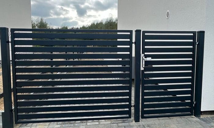 Metal Hegn – JALOUSI portene, med et slankt design og udført i sort pulverlak, står op ad en hvid stukvæg. Disse vedligeholdelsesfrie porte integreres i scenen med træer under en overskyet himmel med udsigt over grå stenbelægning.