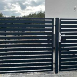 Den moderne, slanke Metal Hegn – JALOUSI hegn og låge med vandrette lameller blander sig stilfuldt på den stenbelagte indkørsel med en hvid bygning sat mod en overskyet himmel som baggrund.