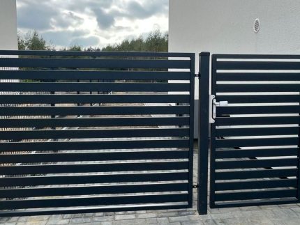 Den moderne, slanke Metal Hegn – JALOUSI hegn og låge med vandrette lameller blander sig stilfuldt på den stenbelagte indkørsel med en hvid bygning sat mod en overskyet himmel som baggrund.