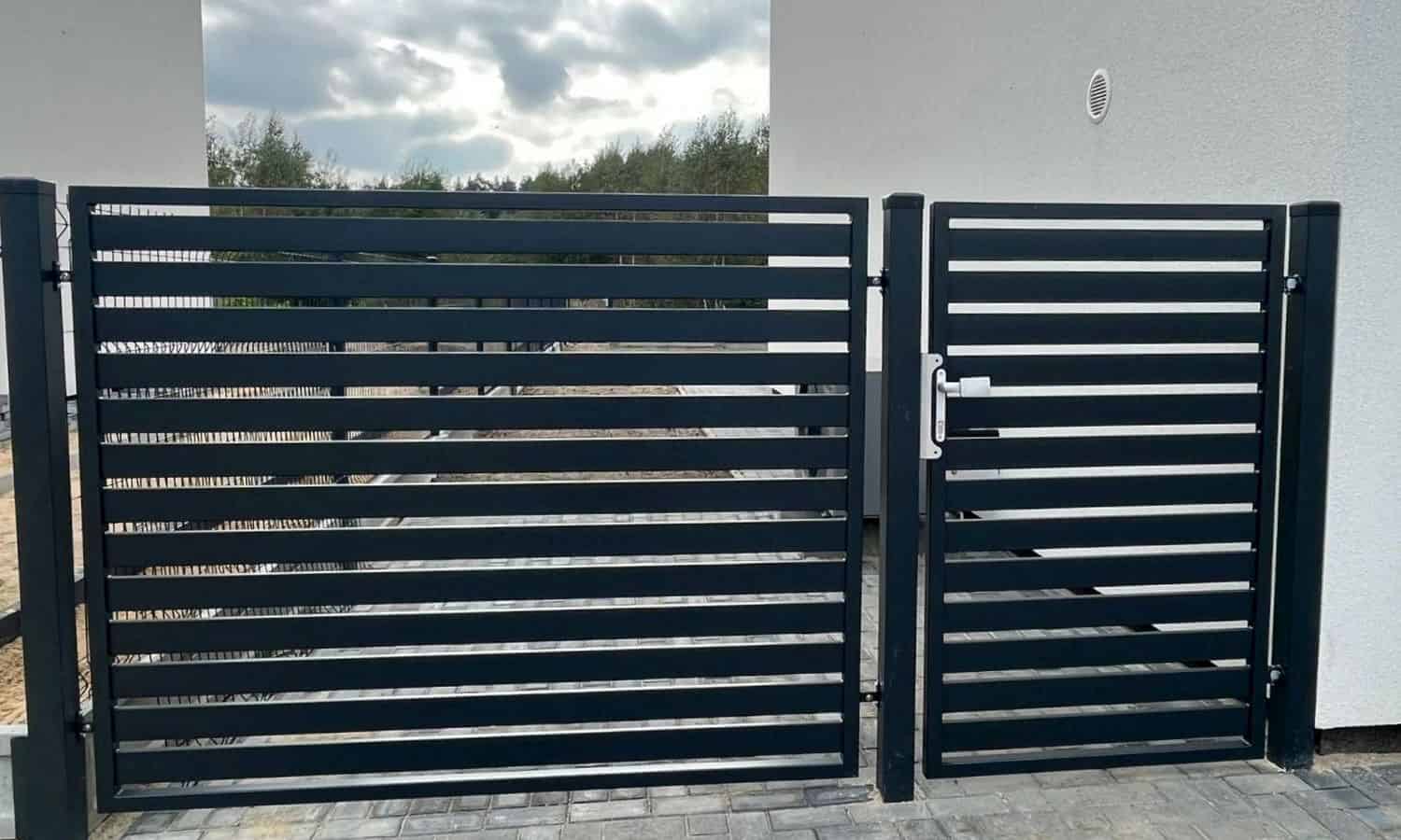 Den moderne, slanke Metal Hegn – JALOUSI hegn og låge med vandrette lameller blander sig stilfuldt på den stenbelagte indkørsel med en hvid bygning sat mod en overskyet himmel som baggrund.