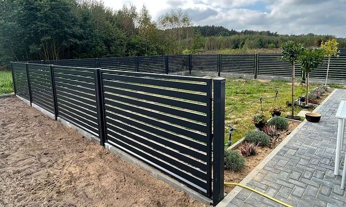 Baghaven, der byder på en Metal Hegn – JALOUSI i et slankt, moderne vandret lameldesign, kan prale af Pulverlakeret-elegance. En sti til højre er flankeret af små planter og buske, mens græs dækker det lukkede område. Træer er synlige mod en overskyet himmel i baggrunden.