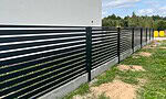 En slank Metal Hegn – JALOUSI, i moderne sort med vandrette lameller, løber langs en bygning på betonfod. Det er pulverlakeret og vedligeholdelsesfrit. Græs, pletter af snavs, træer og en klar himmel ligger ved siden af.