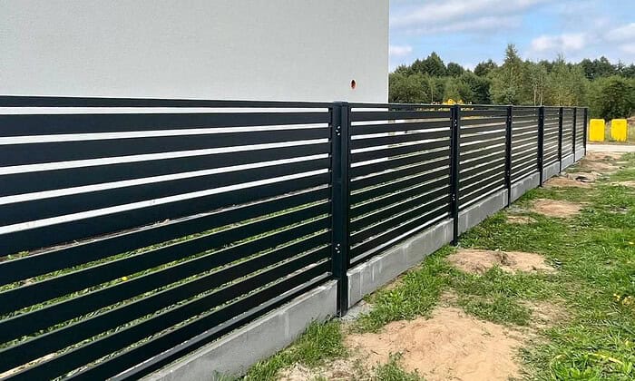 En slank Metal Hegn – JALOUSI, i moderne sort med vandrette lameller, løber langs en bygning på betonfod. Det er pulverlakeret og vedligeholdelsesfrit. Græs, pletter af snavs, træer og en klar himmel ligger ved siden af.