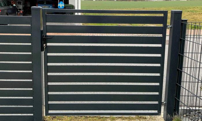 En slank sort Metal Hegn, JALOUSI stil, med vandrette lameller delvist åbne, er fastgjort til et vedligeholdelsesfrit mesh hegn, der fører til en indkørsel med en parkeret bil ved siden af en frodig græsmark.