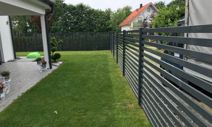 En moderne baghave viser en grøn græsplæne omkranset af Metal Hegn – JALOUSI | Pulverlakeret & Vedligeholdelsesfrit med slanke vandrette lameller. En lille gårdhave med potteplanter og en hvid stol sidder til venstre, mens træer og et hvidt hus med rødt tag fuldender scenen.