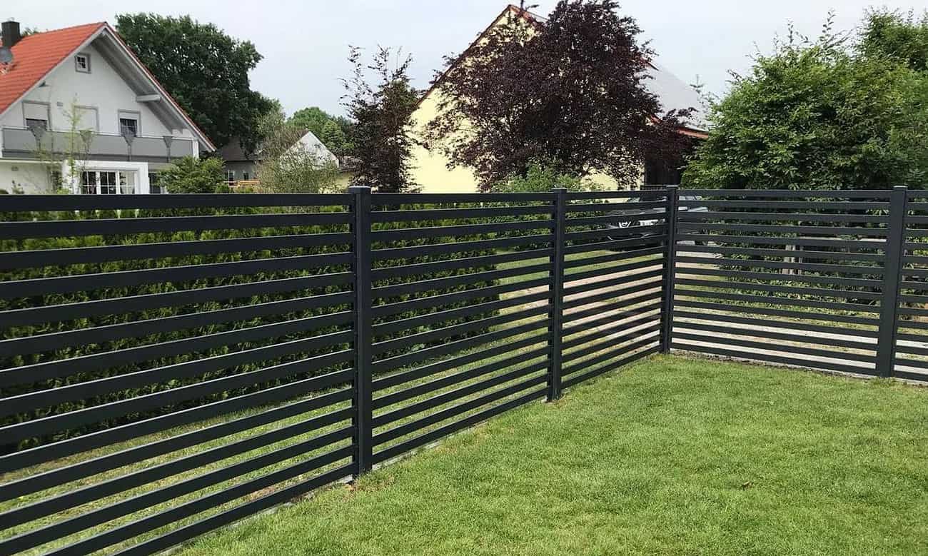En Metal Hegn – JALOUSI med vandrette lameller omslutter en frodig græsplæne, indrammet af træer og buske. Bag vedligeholdelsesfrit hegn ses et gult gavltagshus og et hvidt hus med rødt tag.
