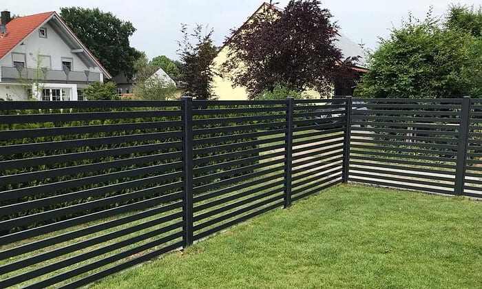 En Metal Hegn – JALOUSI med vandrette lameller omslutter en frodig græsplæne, indrammet af træer og buske. Bag vedligeholdelsesfrit hegn ses et gult gavltagshus og et hvidt hus med rødt tag.