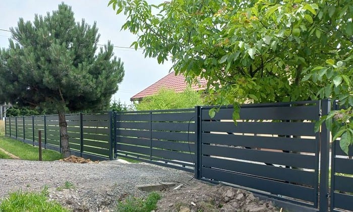 Et slankt, moderne sort Metal Hegn – JALOUSI hegn med vandrette lameller sidder nær et forstadshus med rødt tag, omgivet af frodige grønne områder. Denne vedligeholdelsesfrit metal hegn løber langs en grusbane, og tilbyder holdbarhed på grund af dens pulverlakeret finish.