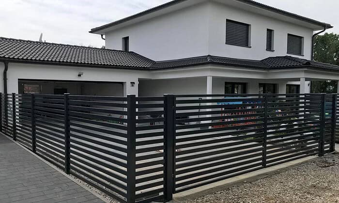 Et moderne hvidt to-etagers hus med mørkt tag har vandrette JALOUSI vinduesskodder og er omgivet af en slank vedligeholdelsesfrit Metal Hegn – JALOUSI. Til ejendommen hører brolagt indkørsel og synlig have bag hegnet.