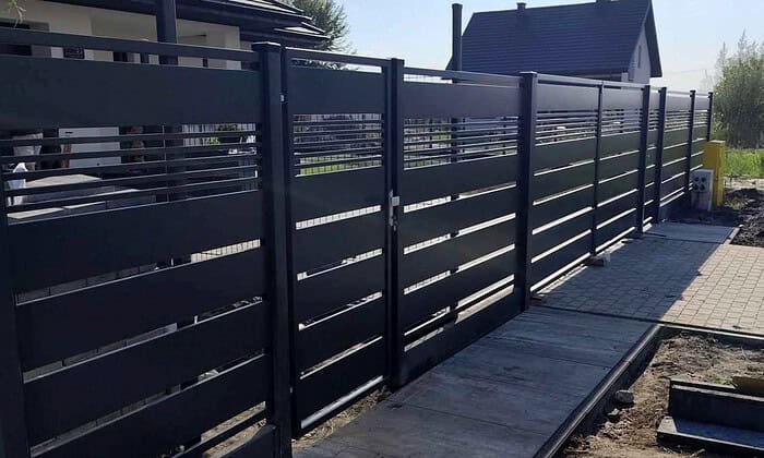 Det moderne boligområde har en moderne sort "Metal Hegn – JALOUSI" med vandrette lameller langs en asfalteret gangsti. Dette vedligeholdelsesfrit hegn er pulverlakeret for holdbarhed, samtidig med at det giver privatliv og stil.