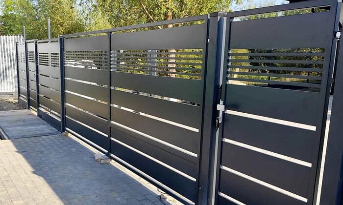 Metal Hegn – JALOUSI er et stilfuldt, vedligeholdelsesfrit metalhegn med vandrette lameller, der giver et glimt af vejen forbi. Dens moderne design sidder elegant på en brolagt overflade, indrammet af frodige grønne områder i baggrunden.