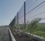 Et højt metaltrådshegn løber langs en asfalteret vej, der ligner det elegante design af højkvalitets hegn og låger. Buske og blomstrende planter vokser ved siden af hegnet, med en klar blå himmel i baggrunden.