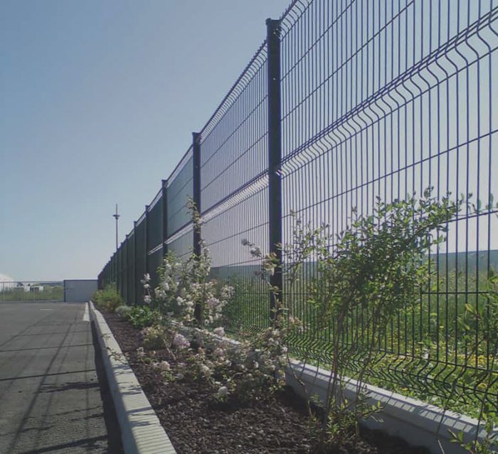 Et højt metaltrådshegn løber langs en asfalteret vej, der ligner det elegante design af højkvalitets hegn og låger. Buske og blomstrende planter vokser ved siden af hegnet, med en klar blå himmel i baggrunden.