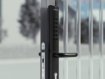 En Locinox VERA sort elektronisk portlås med tastatur og håndtag er monteret på en metallåge, sat på baggrund af slørede huse og løv, ideel til at sikre låger og porte.