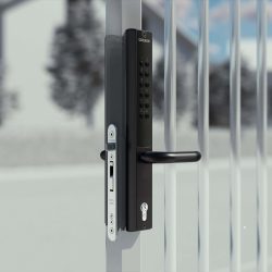 En Locinox VERA sort elektronisk portlås med tastatur og håndtag er monteret på en metallåge, sat på baggrund af slørede huse og løv, ideel til at sikre låger og porte.