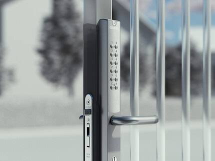 Nærbillede af en moderne metalportlås med Locinox VERA Batteridrevet Kodelås til Låger og Porte, med et tastatur til kodeindtastning og manuel nøgleadgang, sat mod en sløret udendørs baggrund.