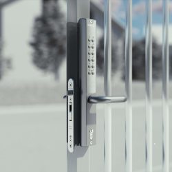 Nærbillede af en moderne metalportlås med Locinox VERA Batteridrevet Kodelås til Låger og Porte, med et tastatur til kodeindtastning og manuel nøgleadgang, sat mod en sløret udendørs baggrund.