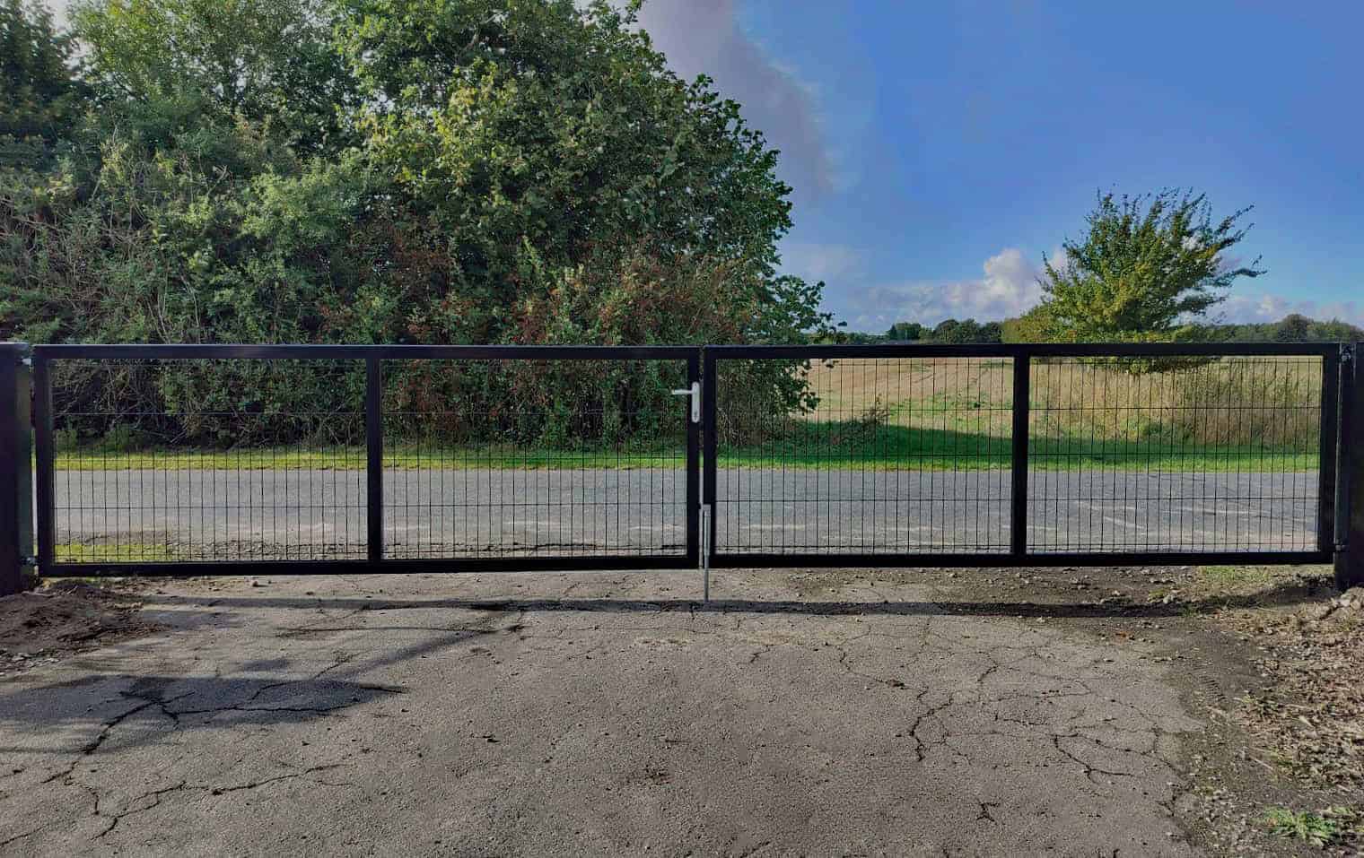 En sort metalhegnslåge står solidt lukket hen over den revnede asfaltindkørsel. Bagved grænser træer og buske en græsmark under en blå himmel med spredte skyer, hvilket skaber en malerisk forside, der indbyder til nysgerrighed.