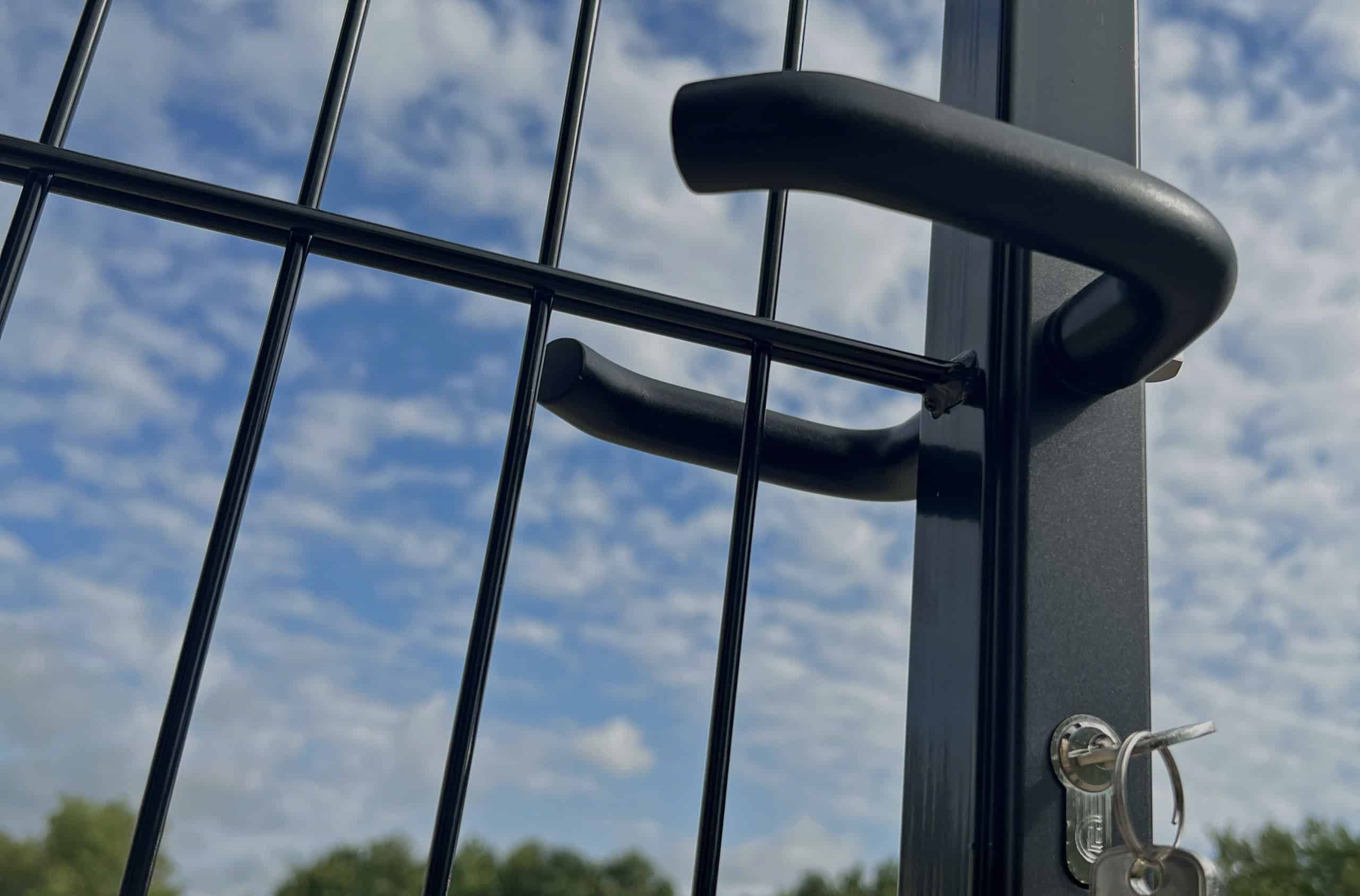 Et nærbillede af en sort metalport med håndtag og lås, stående mod en overskyet blå himmel og fjerne grønne træer. Porten, der står lidt på klem med nøgler indsat i låsen, tjener som en tavs vogter på denne fredfyldte forside.