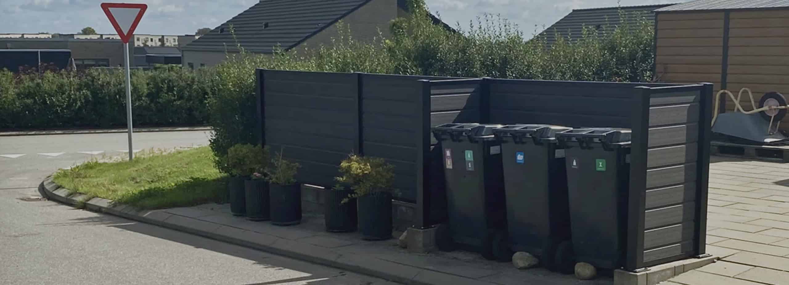 Fire sorte skraldespande på hjul står opstillet ved siden af en lækker komposithegn på et fortovshjørne. Potteplanter placeres ved siden af hegnet og tilføjer et strejf af grønt. Et vigeskilt er synligt på gaden, og huse danner baggrunden.