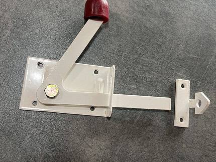 En Lukkebeslag Hvid Metal lås med rødt håndtag, monteret på en plan overflade, passer til trælåger. Det inkluderer en vægtstangsarm på en rektangulær base med skruer og et ekstra lille metalstykke, der sandsynligvis er en del af systemet.