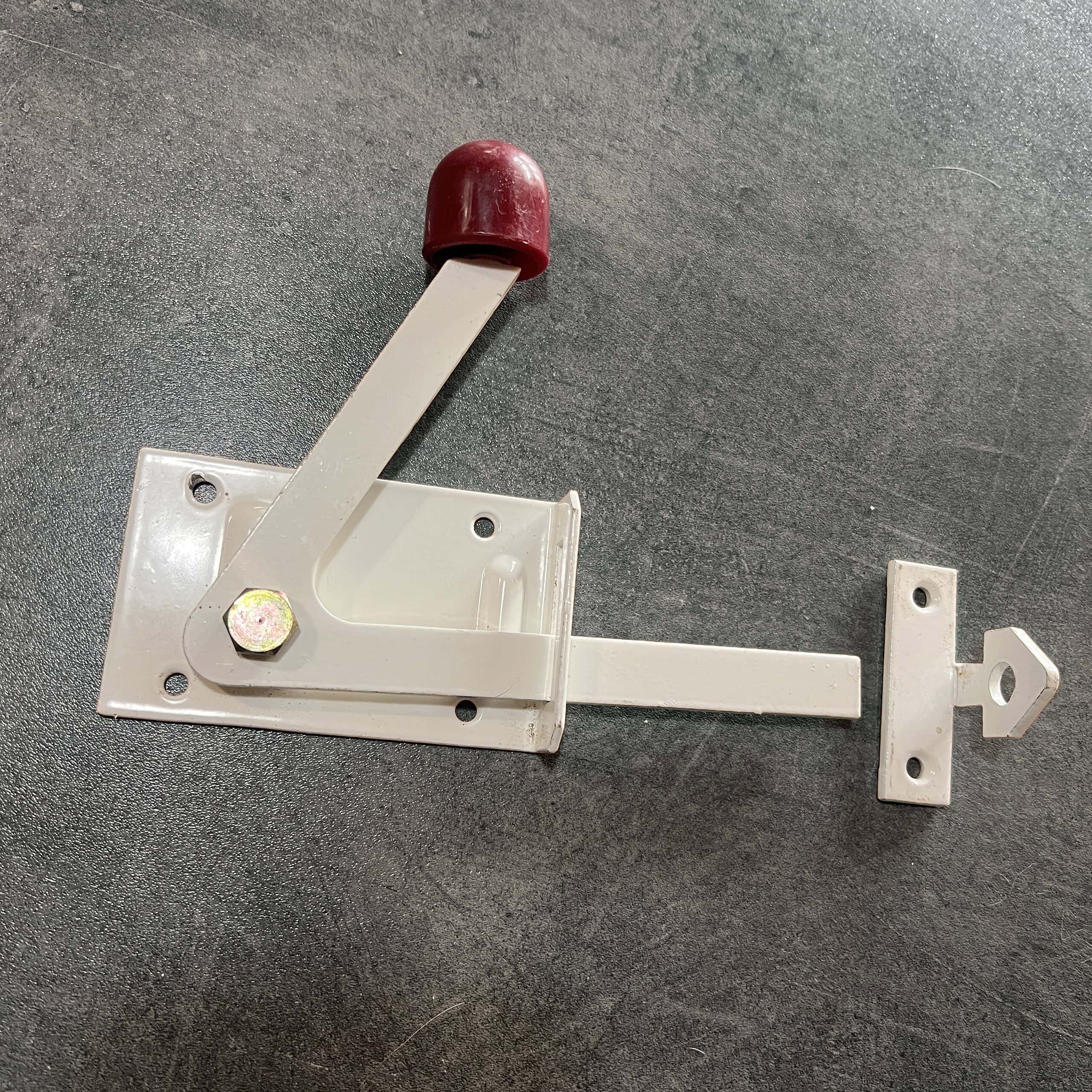 En Lukkebeslag Hvid Metal lås med rødt håndtag, monteret på en plan overflade, passer til trælåger. Det inkluderer en vægtstangsarm på en rektangulær base med skruer og et ekstra lille metalstykke, der sandsynligvis er en del af systemet.