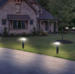 Et murstenshus med skrånende tag er omgivet af en velholdt græsplæne. Moderne Stålstolpe pulverlakeret sort på fod inkl. zquare lys udendørslys langs indkørslen og kaster et blødt skær. Et træ er nær fronten, og buske er pænt arrangeret langs facaden.