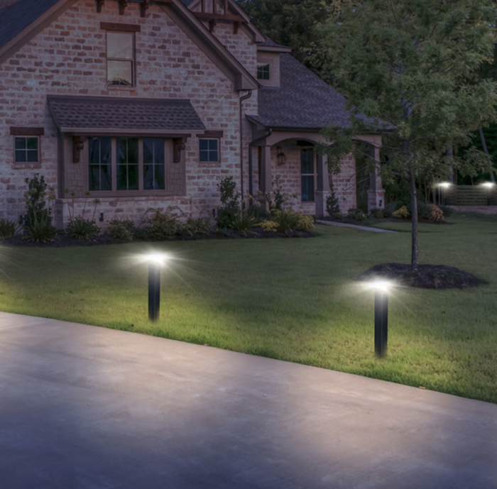 Et murstenshus med skrånende tag er omgivet af en velholdt græsplæne. Moderne Stålstolpe pulverlakeret sort på fod inkl. zquare lys udendørslys langs indkørslen og kaster et blødt skær. Et træ er nær fronten, og buske er pænt arrangeret langs facaden.